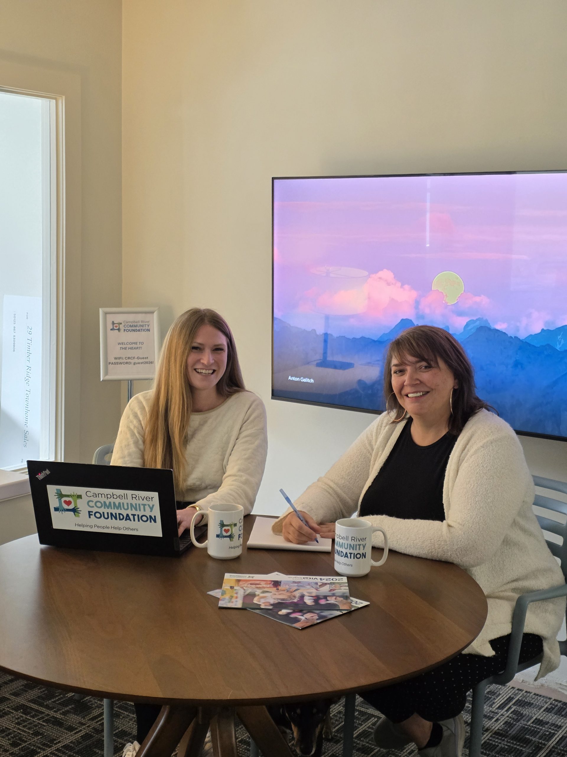 Tech Resident 1 Two people smiling while coworking at a round table with Campbell River Community Foundation branded mugs, laptop, and 2024 Vital Signs reports.